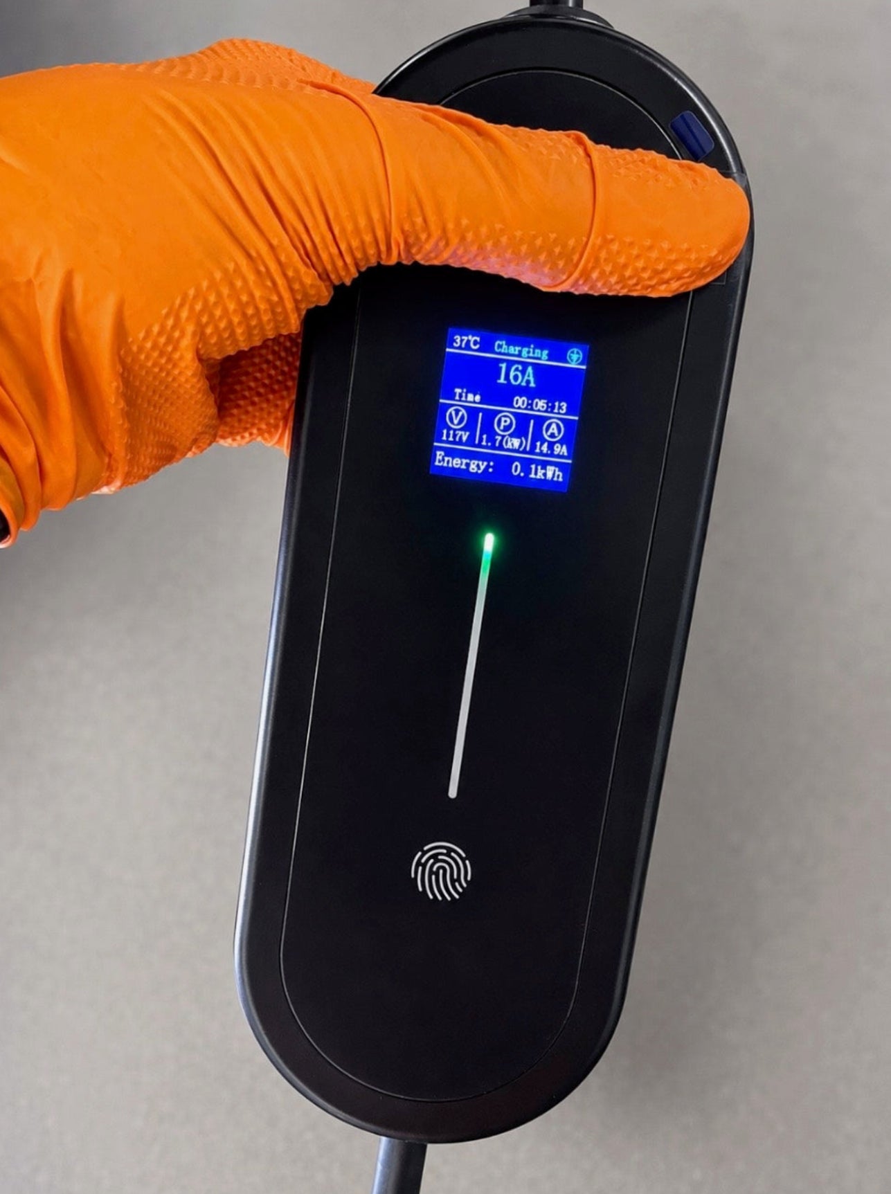 A close-up of a hand wearing an orange textured glove holding a black electronic device with a small blue display screen and a fingerprint sensor, against a neutral gray background.